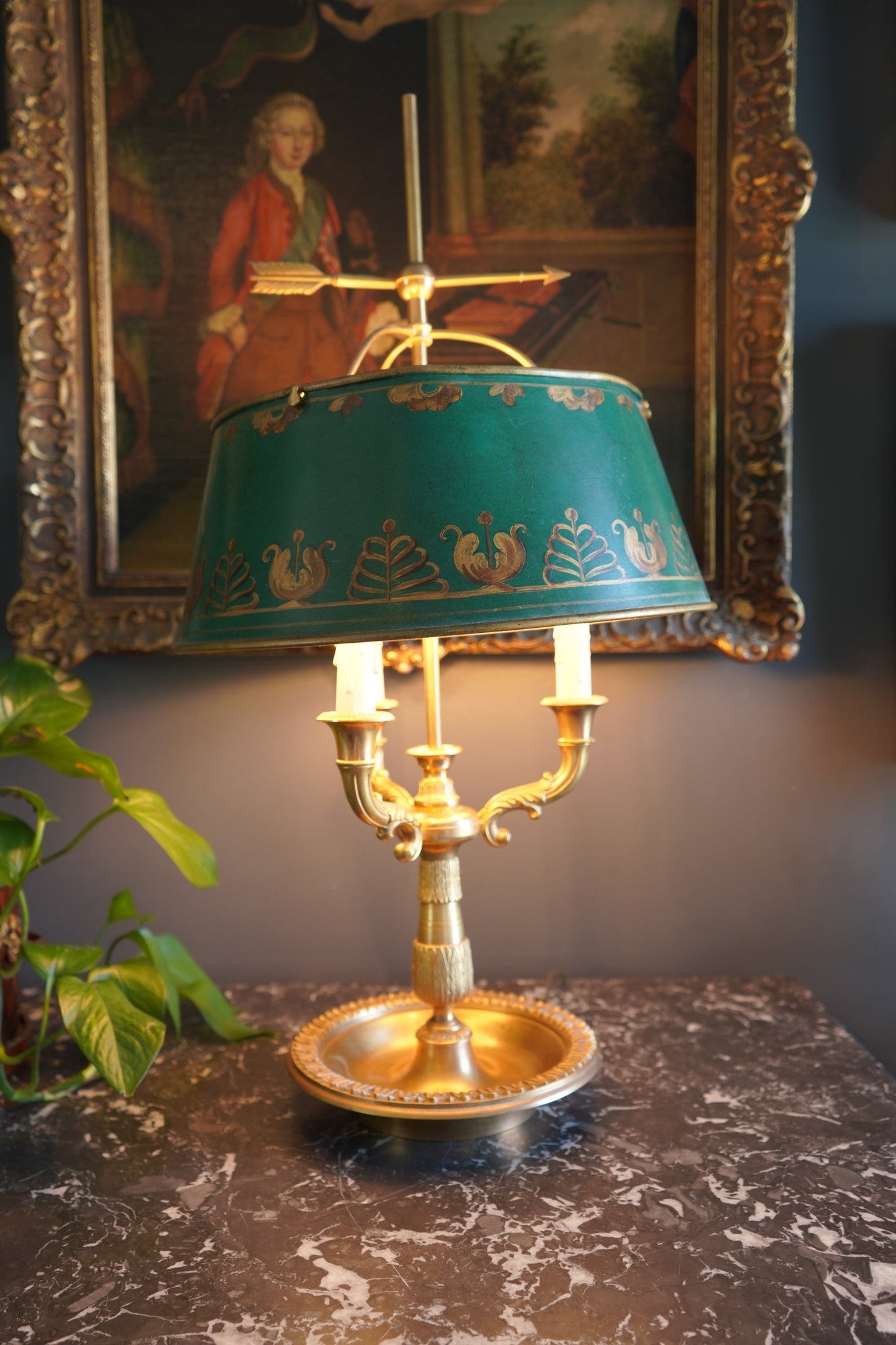 Decorative lamp with green shade and gold base on a dark surface, with a painting in the background.