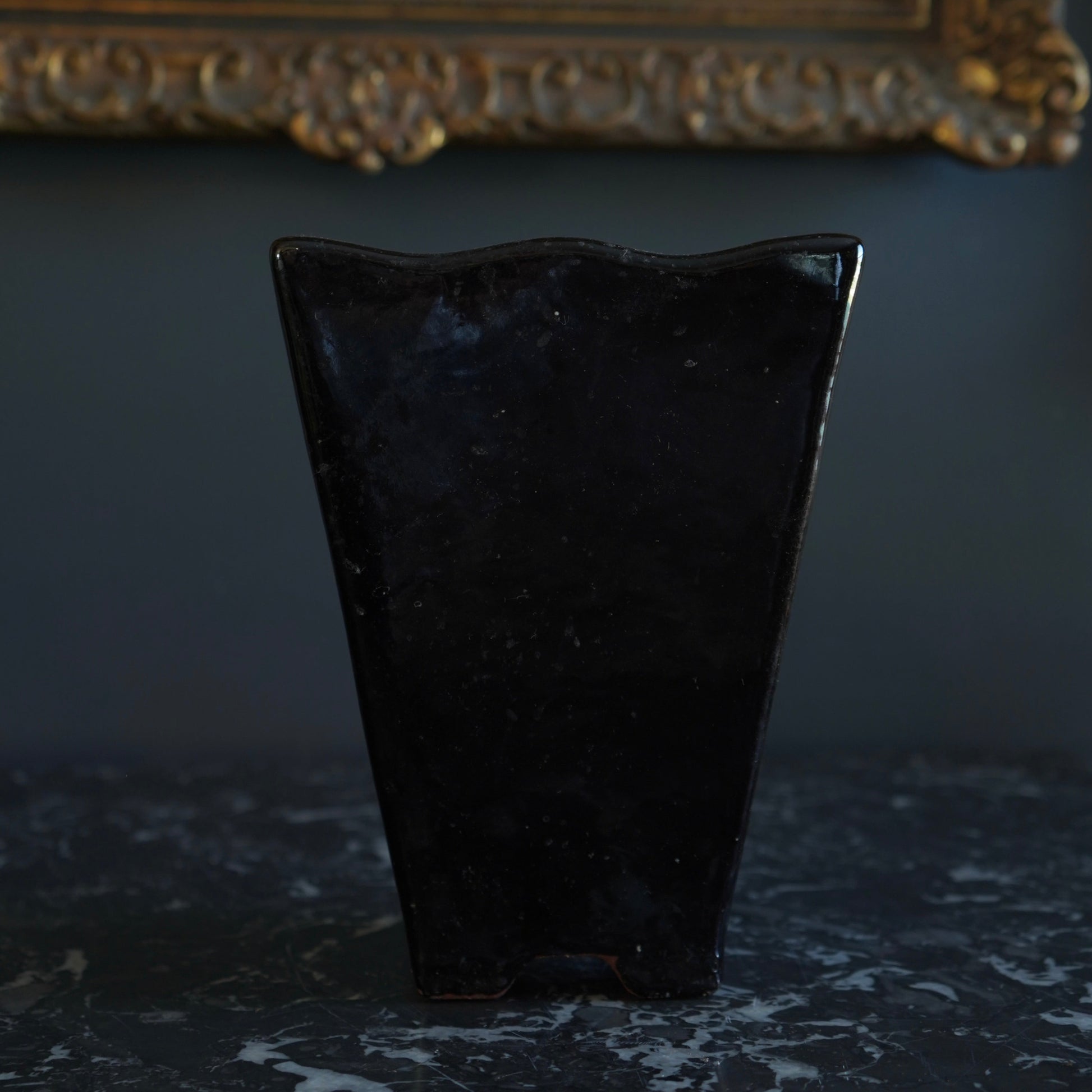 Black vase on a dark surface with a framed picture in the background