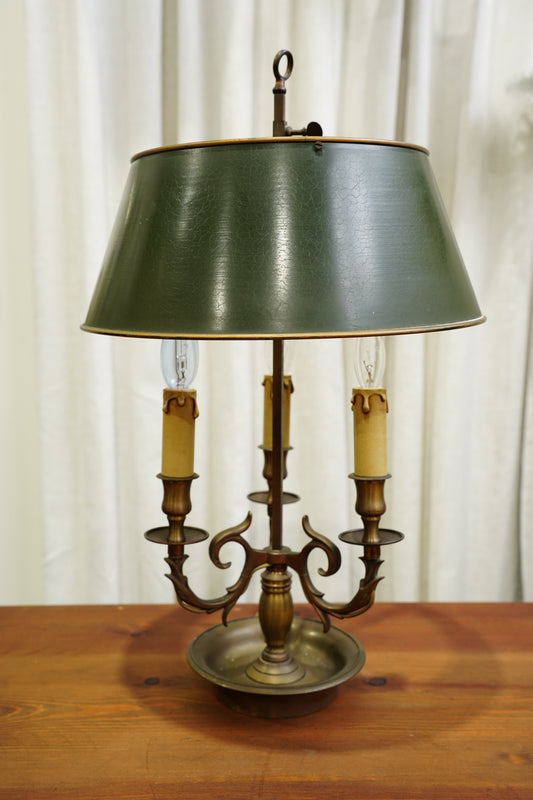 A vintage three-light candelabra table lamp with a dark green metal shade and an antique bronze metal base, displayed on a wooden table against a white curtain backdrop.
