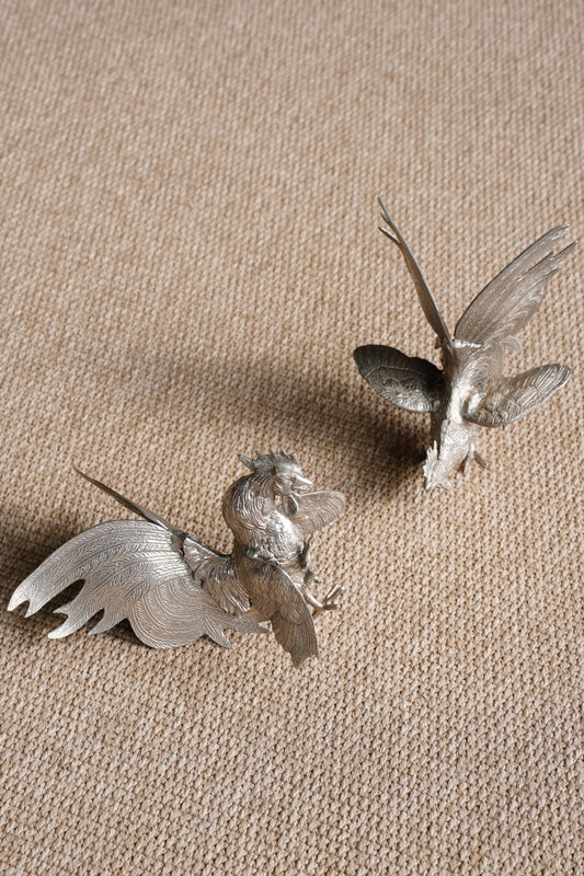 Two small metallic bird figurines with spread wings, placed on a textured beige fabric background.