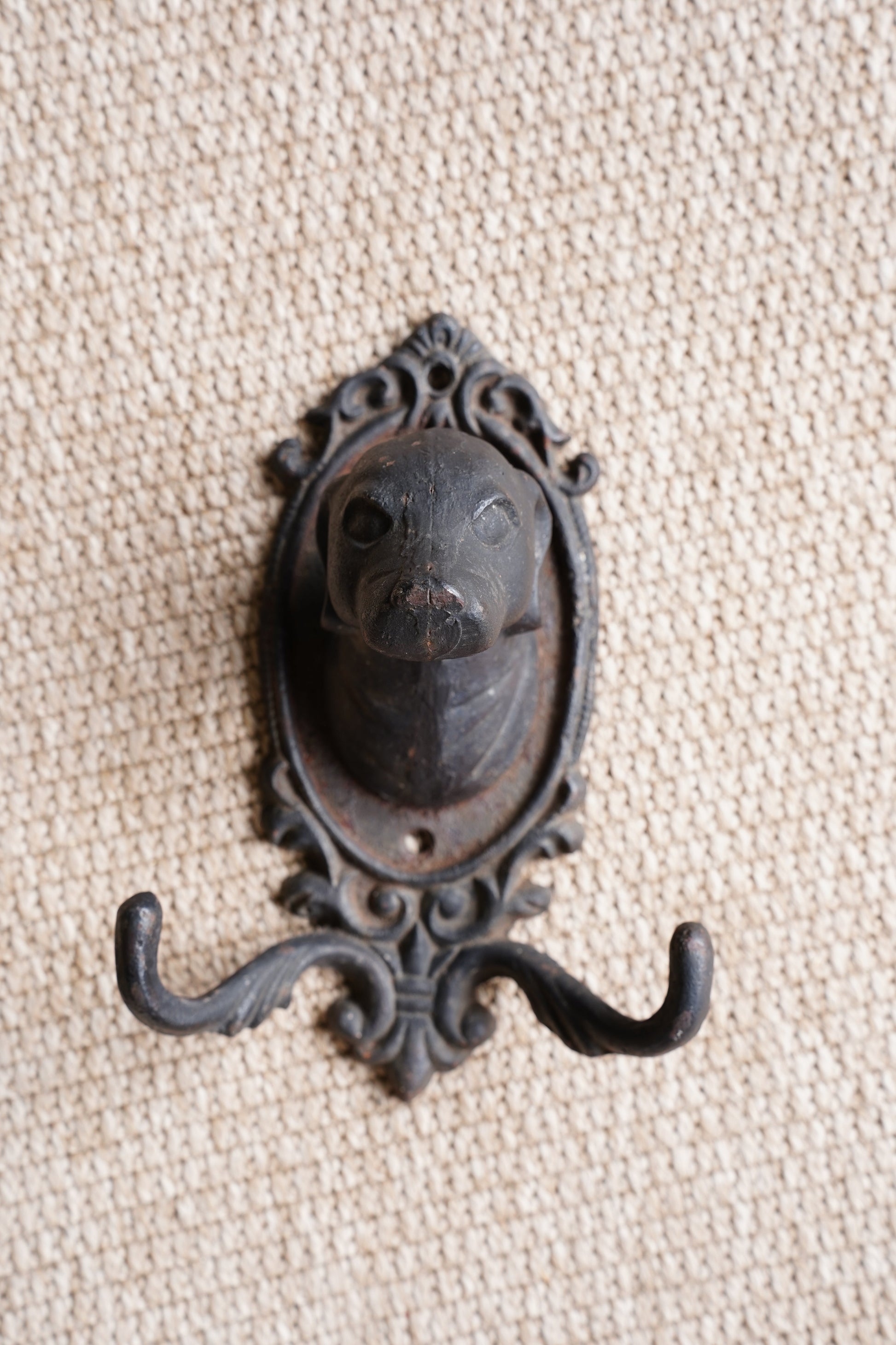 An ornate vintage-style black metal hook with an oval decorative frame, displayed on a textured beige woven fabric background.