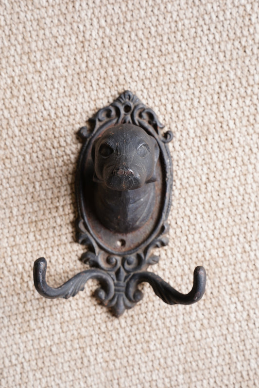 An ornate vintage-style black metal hook with an oval decorative frame, displayed on a textured beige woven fabric background.