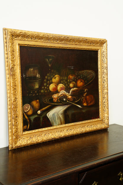 A framed still life oil painting depicting fruit, grapes, and a roasted bird on a table, housed in an ornate gold frame and leaning against a white wall on a dark wooden surface.