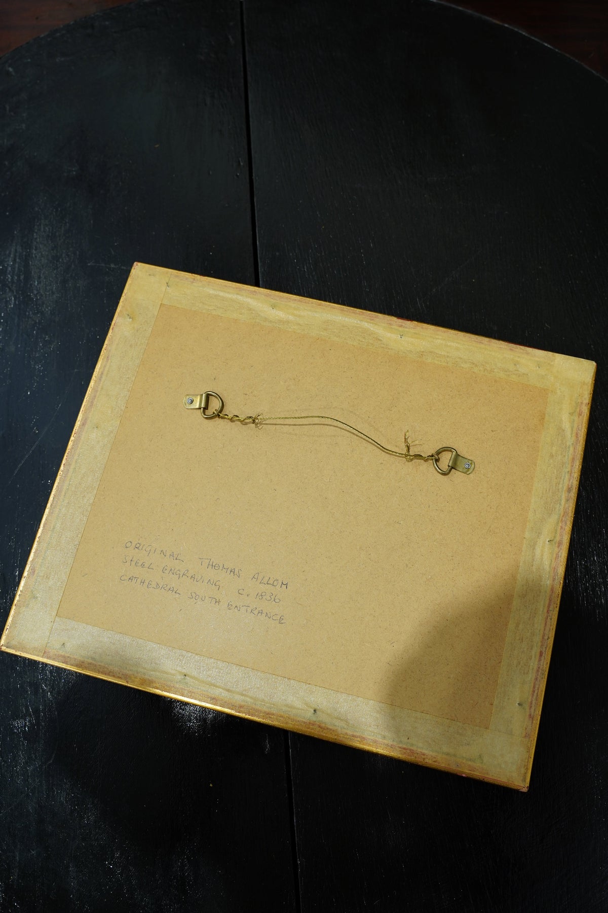 The back of a square wooden picture frame with a light tan backing board, handwritten notes in the top left, and a brass hanging loop and key attached, resting on a dark wooden surface.