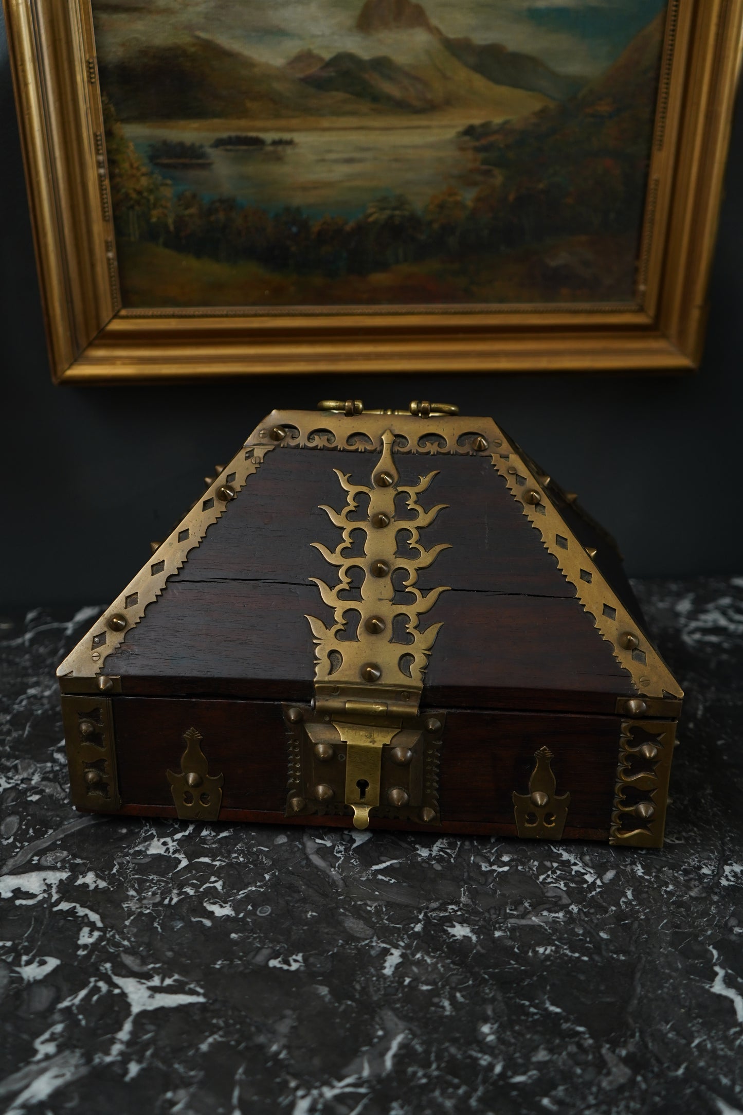 Decorative wooden box with brass hinges on a dark surface next to a framed painting.